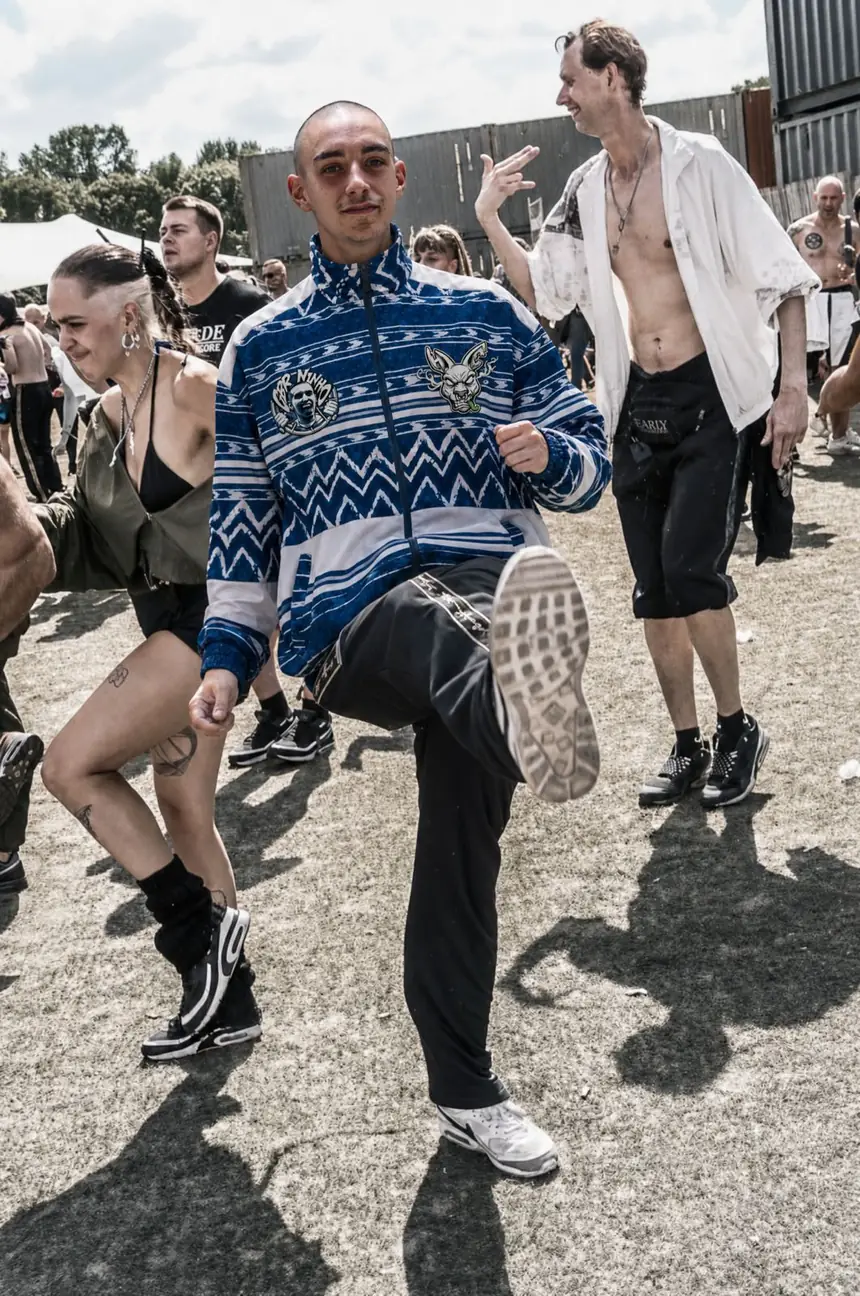 Gabber Ninho dancing hakken in a festival field, knee up, in a blue KANG tracksuit