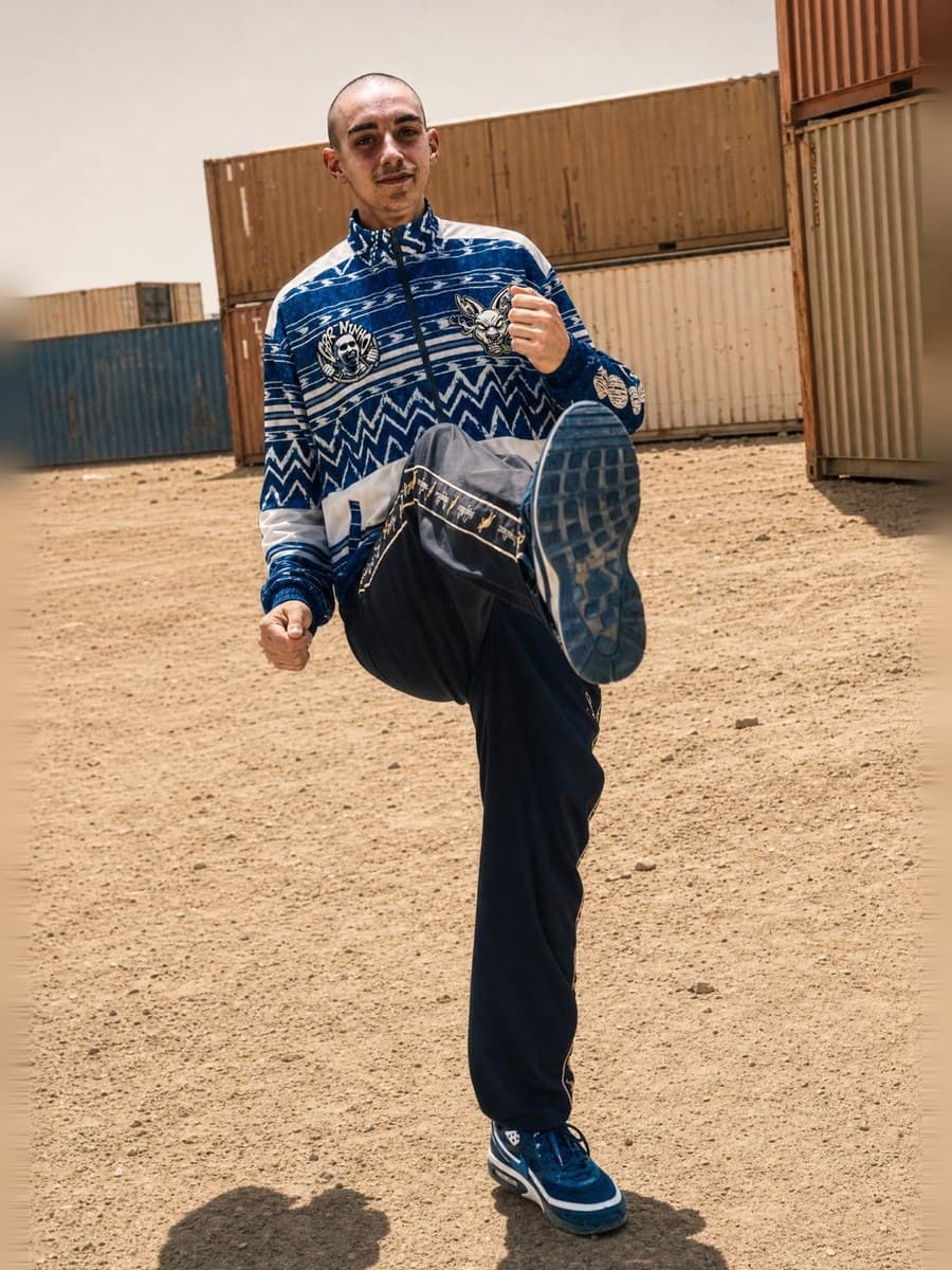 Dancer with a knee raised on a dirt yard, shipping containers behind
